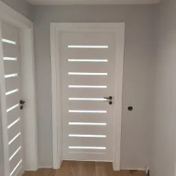 Nowoczesne białe drzwi wewnętrzne z poziomymi przeszkleniami, zamontowane w szarej ścianie nad drewnianą podłogą, widoczna instalacja elektryczna.