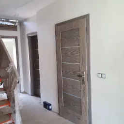 Montaż drzwi wewnętrznych w Pcimiu, widoczne surowe ściany, zabezpieczone schody i pudełko z akcesoriami.