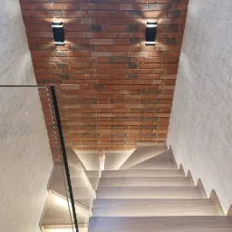 Nowoczesne schody z jasnego drewna z podświetleniem LED, szklaną balustradą i ścianą z czerwonej cegły podświetloną czarnymi lampami ściennymi.
