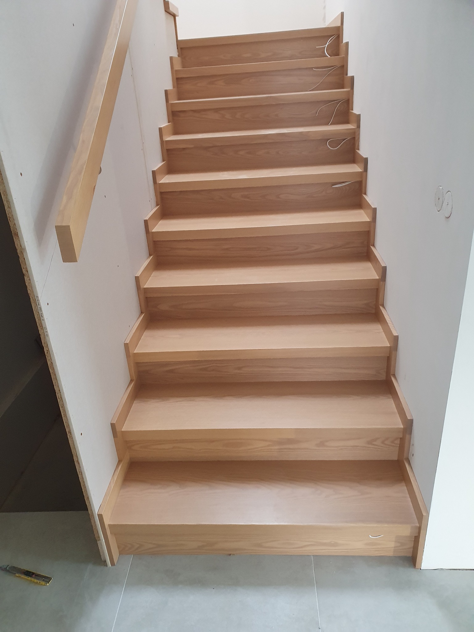 Jasne, drewniane schody z prostymi stopniami, widoczne kable elektryczne wystające spod stopni, przy ścianie z płyt gipsowo-kartonowych, w tle fragment narzędzia pomiarowego.