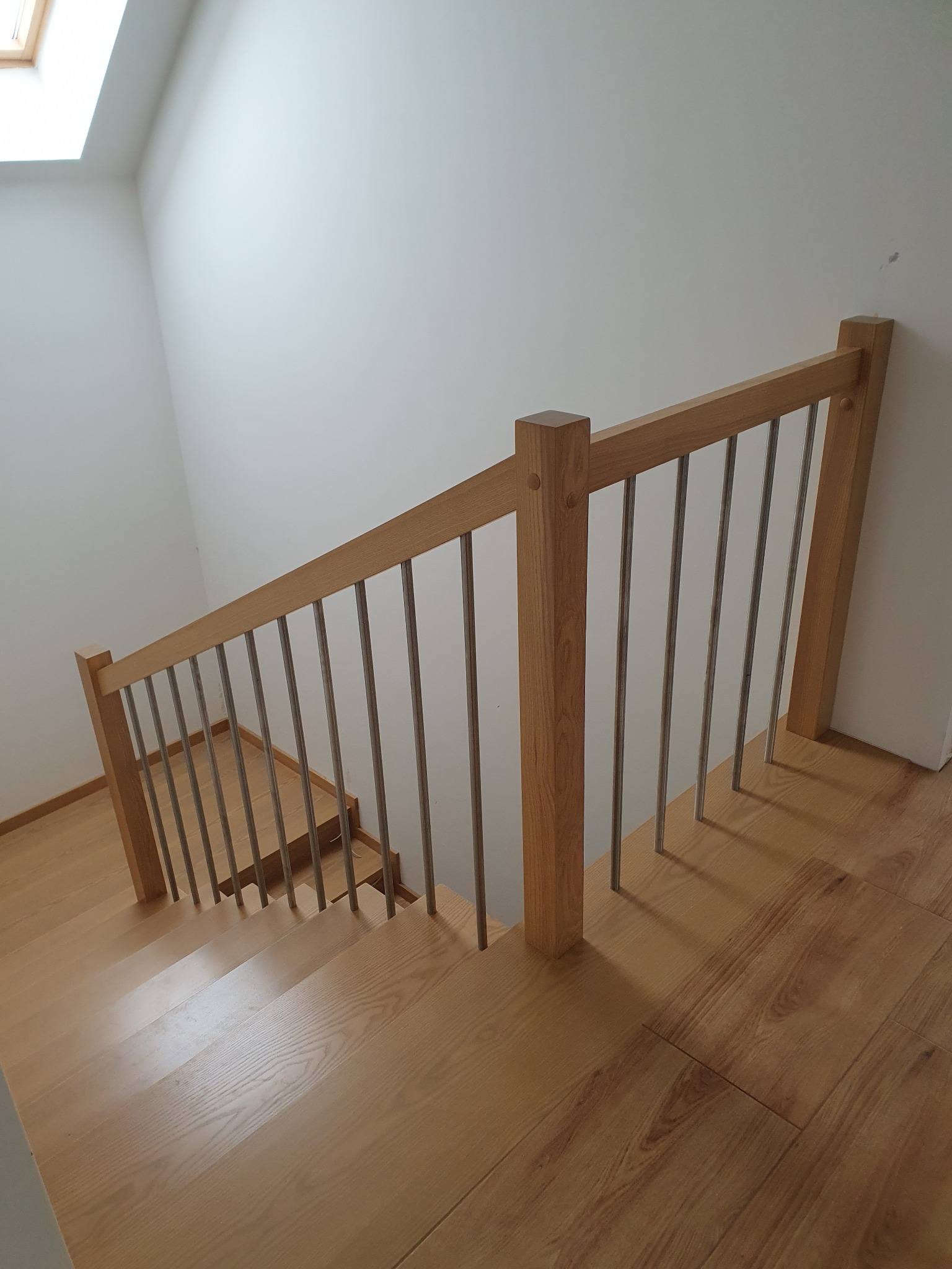 Jasne, drewniane schody z metalowymi tralkami balustrady, widok z góry na dół, naturalne oświetlenie z okna dachowego.