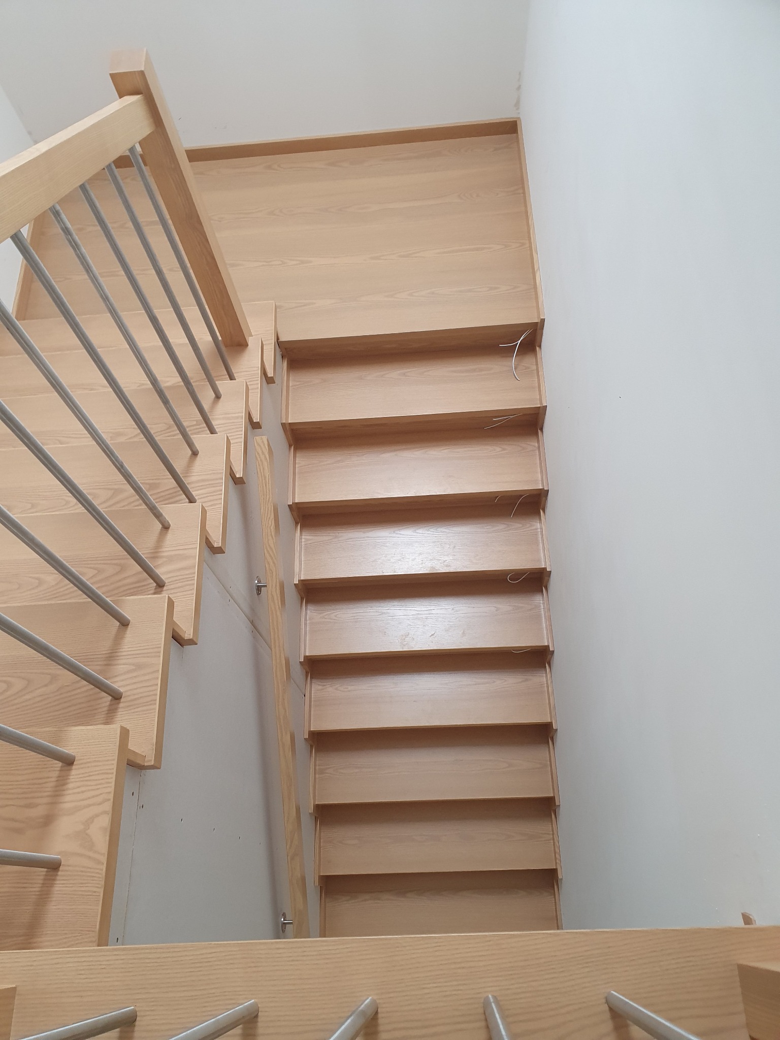 Jasne, drewniane schody z metalowymi poręczami widziane z góry, z widocznymi niedociągnięciami wykończeniowymi na ścianie.