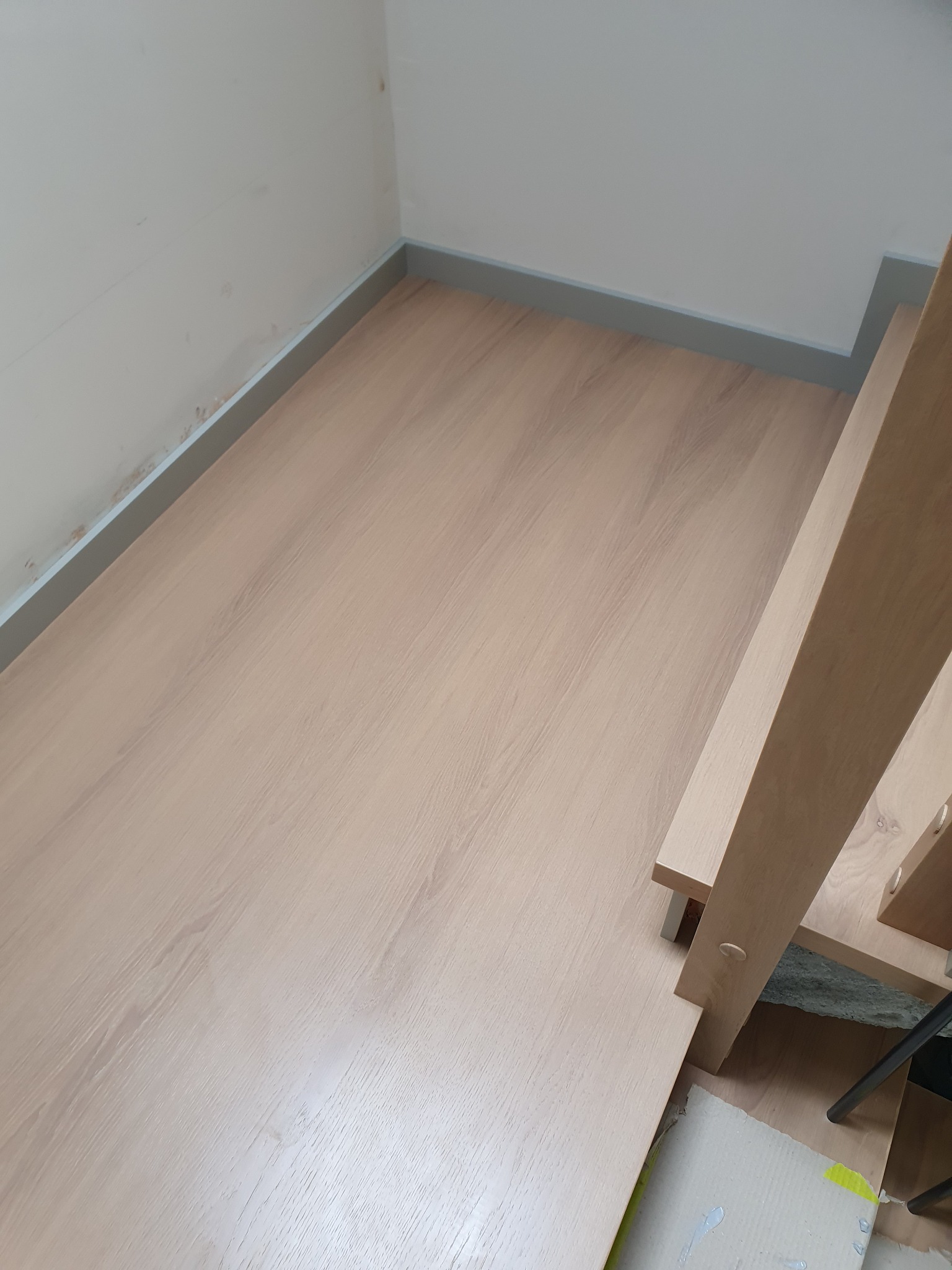 Fragment montażu jasnych schodów z drewnopodobną okleiną, widoczna podłoga i białe ściany z szarą listwą przypodłogową.