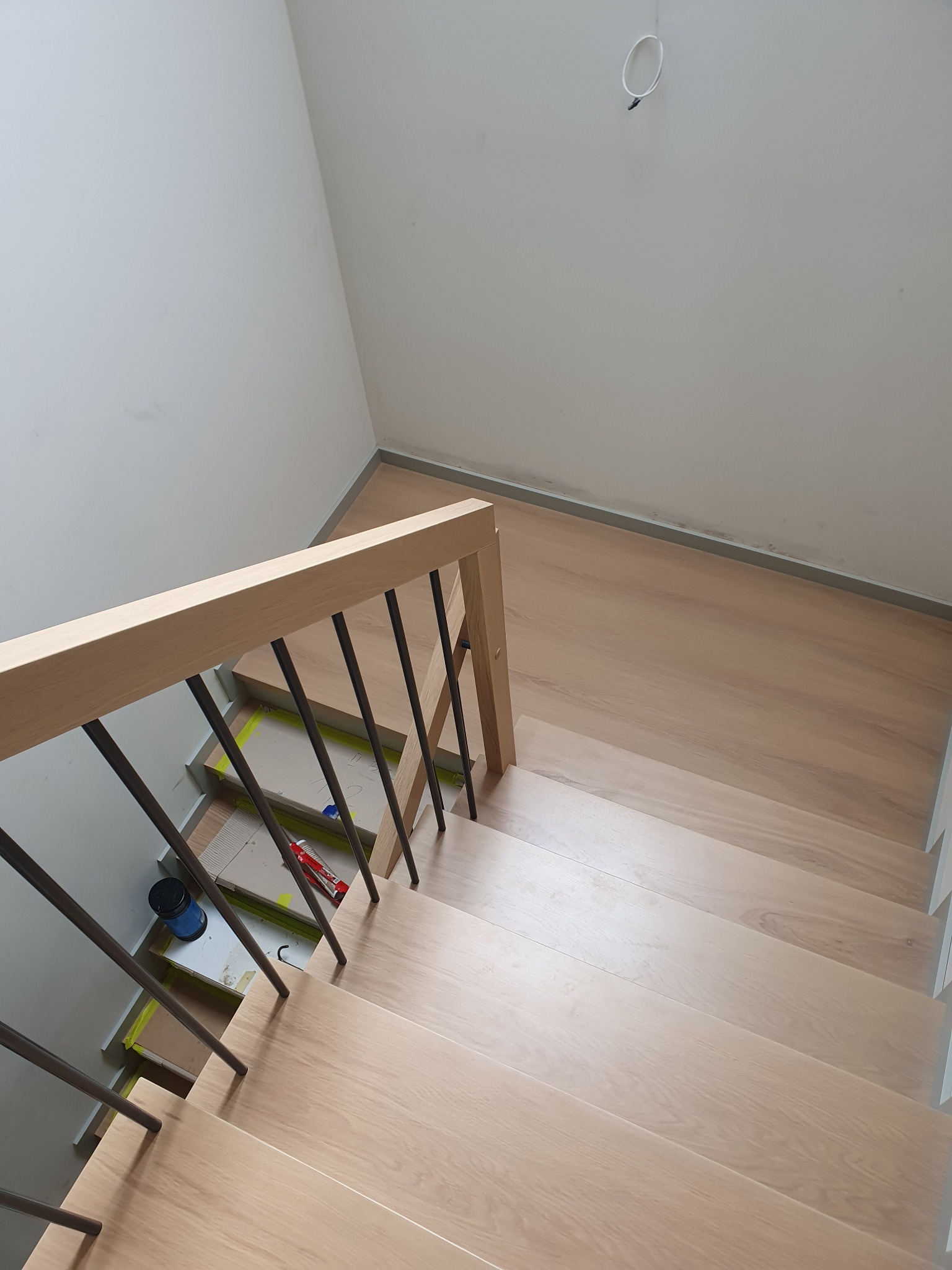 Widok z góry na jasne stopnie z drewnianą balustradą z czarnymi metalowymi prętami, widoczne narzędzia i materiały budowlane na zabezpieczonych schodach.