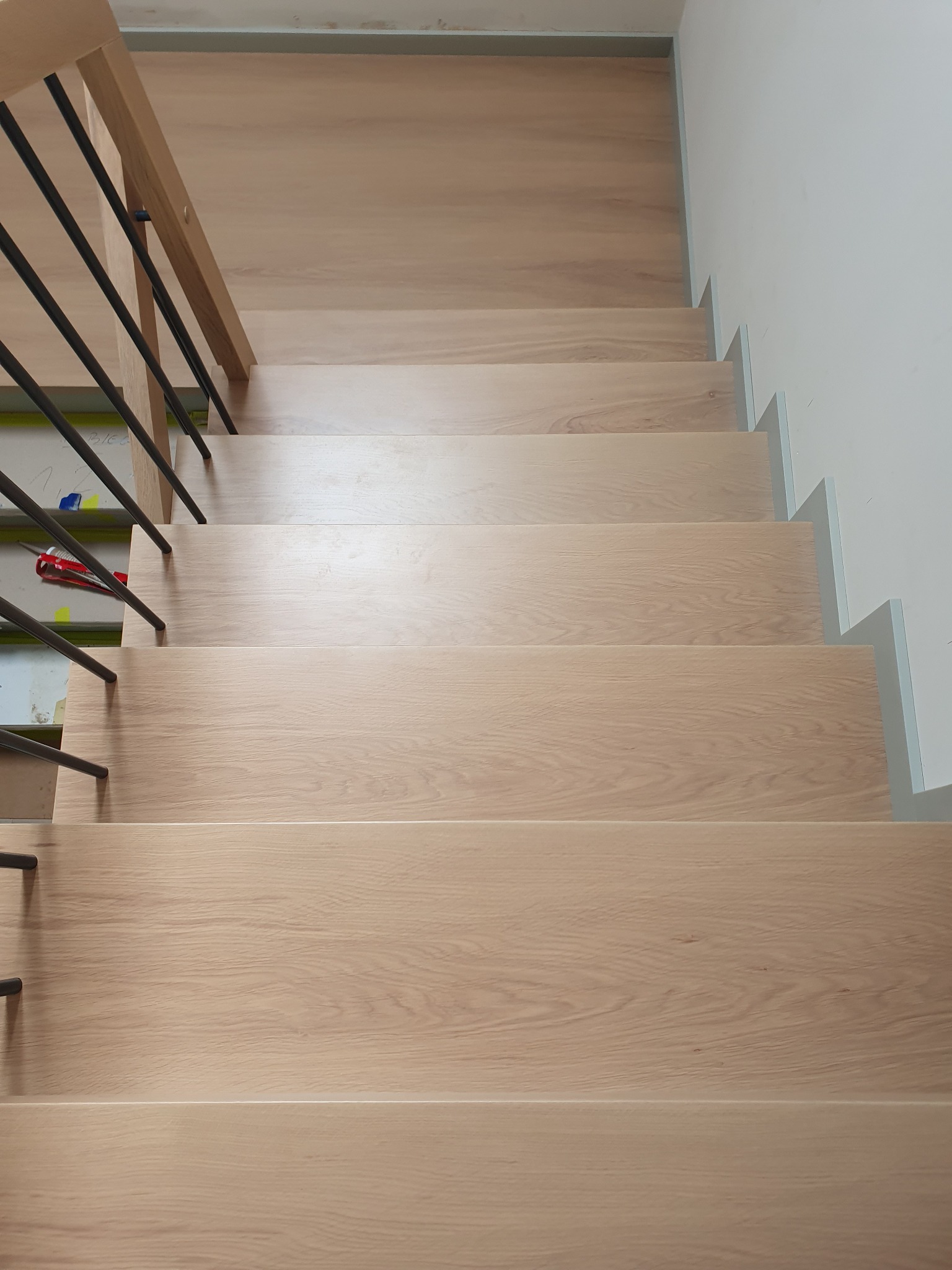 Jasne, drewniane schody z czarnymi, metalowymi balustradami, widok z góry, w tle biała ściana i elementy w trakcie montażu.