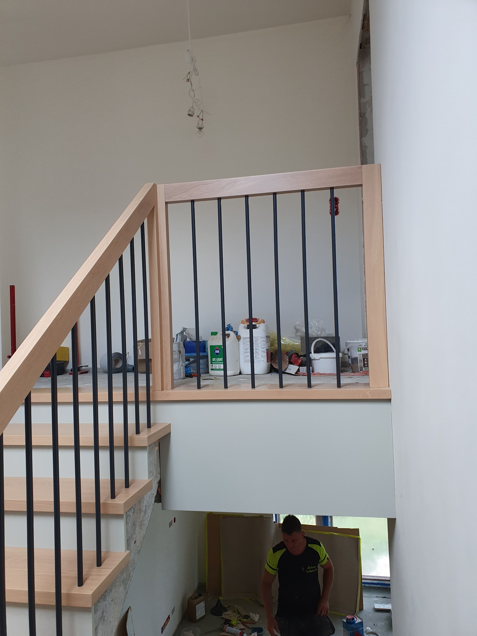 Montaż balustrady z jasnego drewna i czarnych prętów na schodach w surowym wnętrzu, widoczny pracownik w trakcie pracy, narzędzia i materiały budowlane w tle.