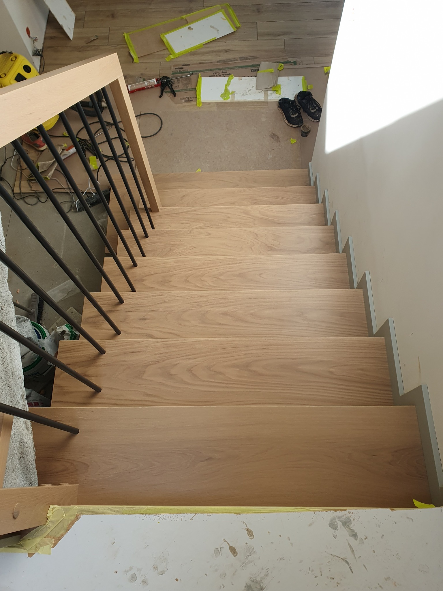 Montaż jasnych, drewnianych stopni schodowych z metalową balustradą, widok z góry na częściowo zabezpieczone folią podłoże i narzędzia pracy.