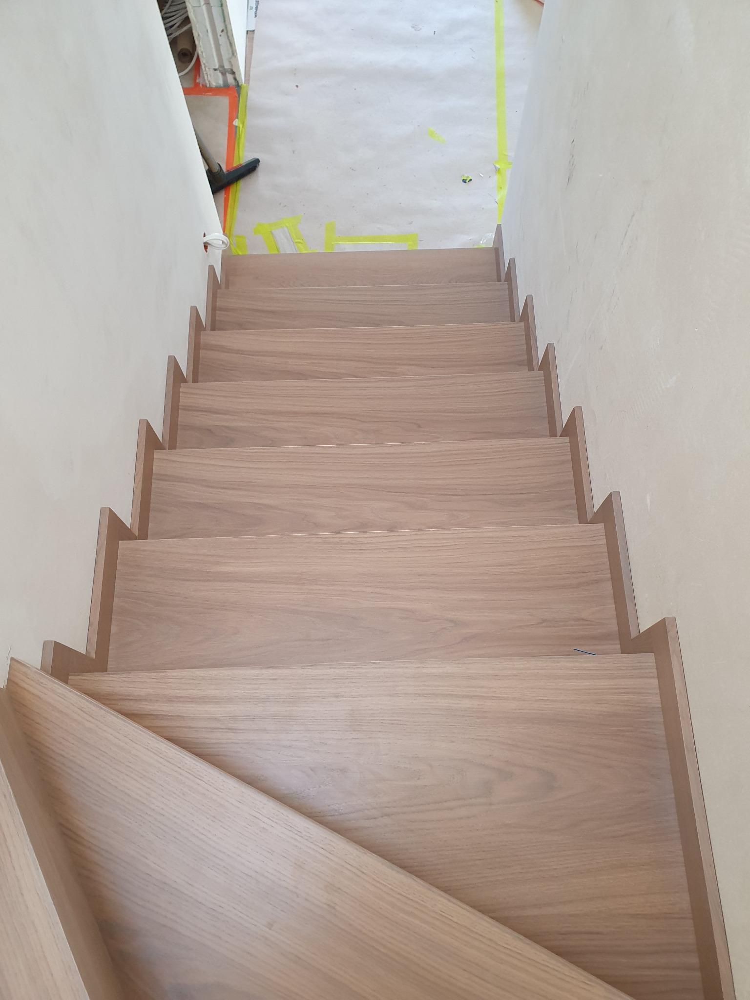 Nowe, jasne stopnie z drewna z widocznym usłojeniem, zamontowane w surowym wnętrzu z żółtą taśmą malarską na podłodze i odkurzaczem w rogu.