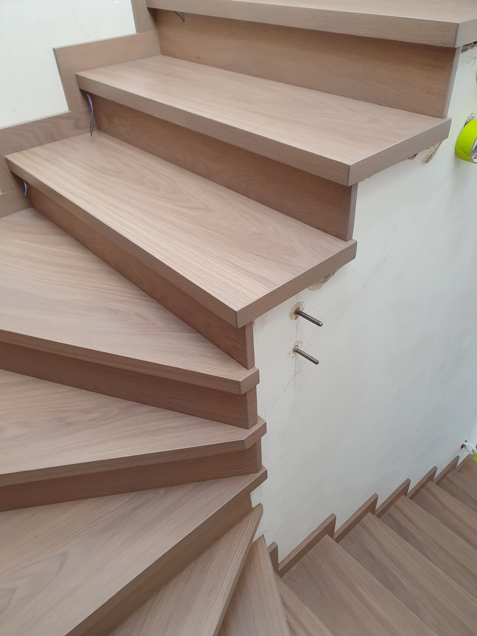 Montaż drewnianych schodów spiralnych z widocznymi metalowymi mocowaniami do ściany i taśmą malarską zabezpieczającą.