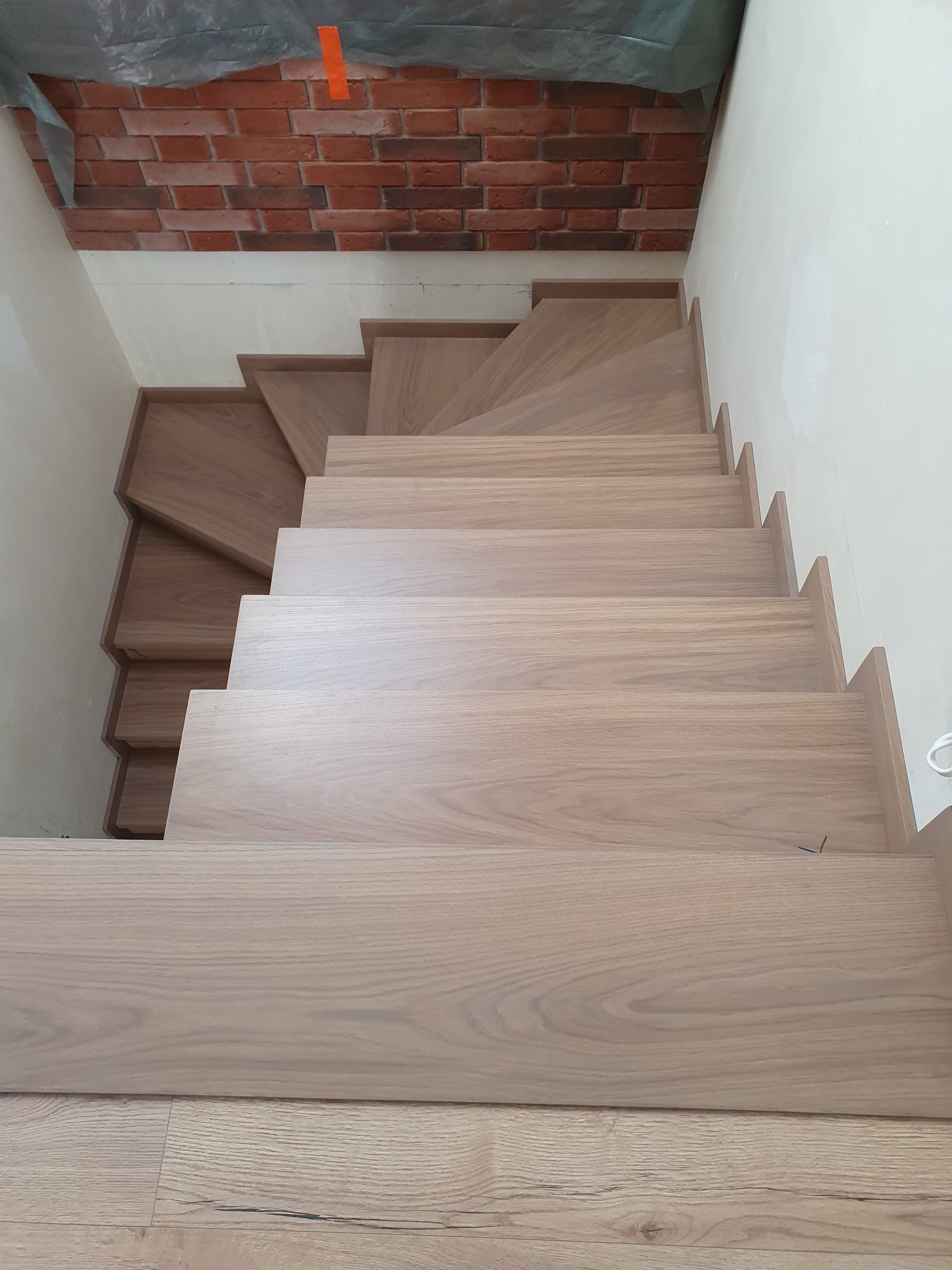 Widok z góry na jasne, drewniane schody z wyraźnym usłojeniem, prowadzące w dół, z fragmentem ceglanej ściany w tle, zabezpieczonej folią malarską.