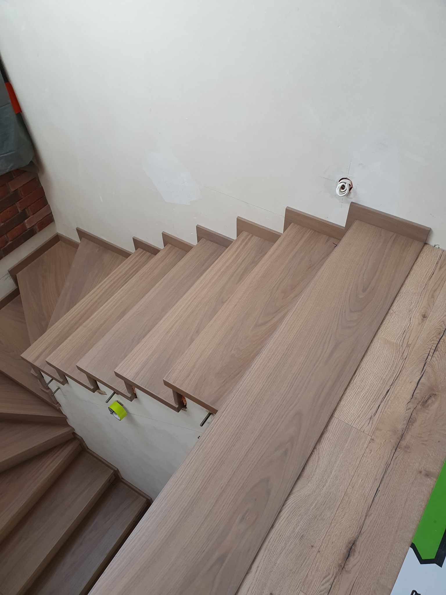 Widok z góry na drewniane stopnie schodów w trakcie montażu, widoczne niedokończone wykończenia ściany i wystające kable elektryczne.