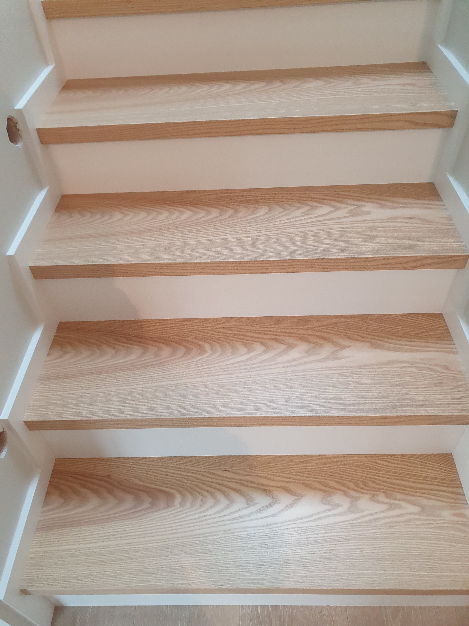 Jasne, drewniane stopnie schodowe z widocznym usłojeniem, osadzone w białej, minimalistycznej konstrukcji; widoczny otwór w bocznej ściance.