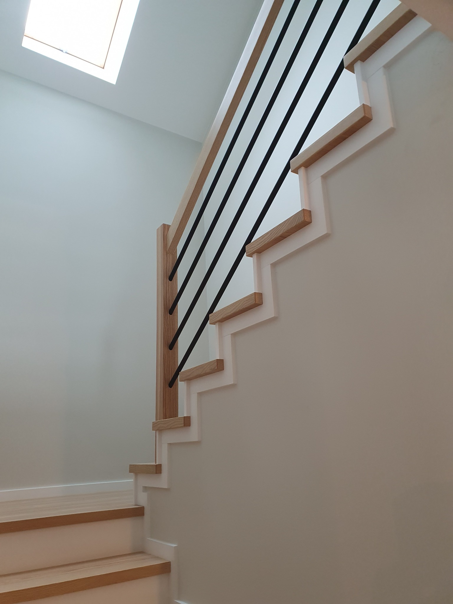 Nowoczesne schody z jasnego drewna z czarnymi, pionowymi balustradami na tle białych ścian i sufitu z oknem dachowym.