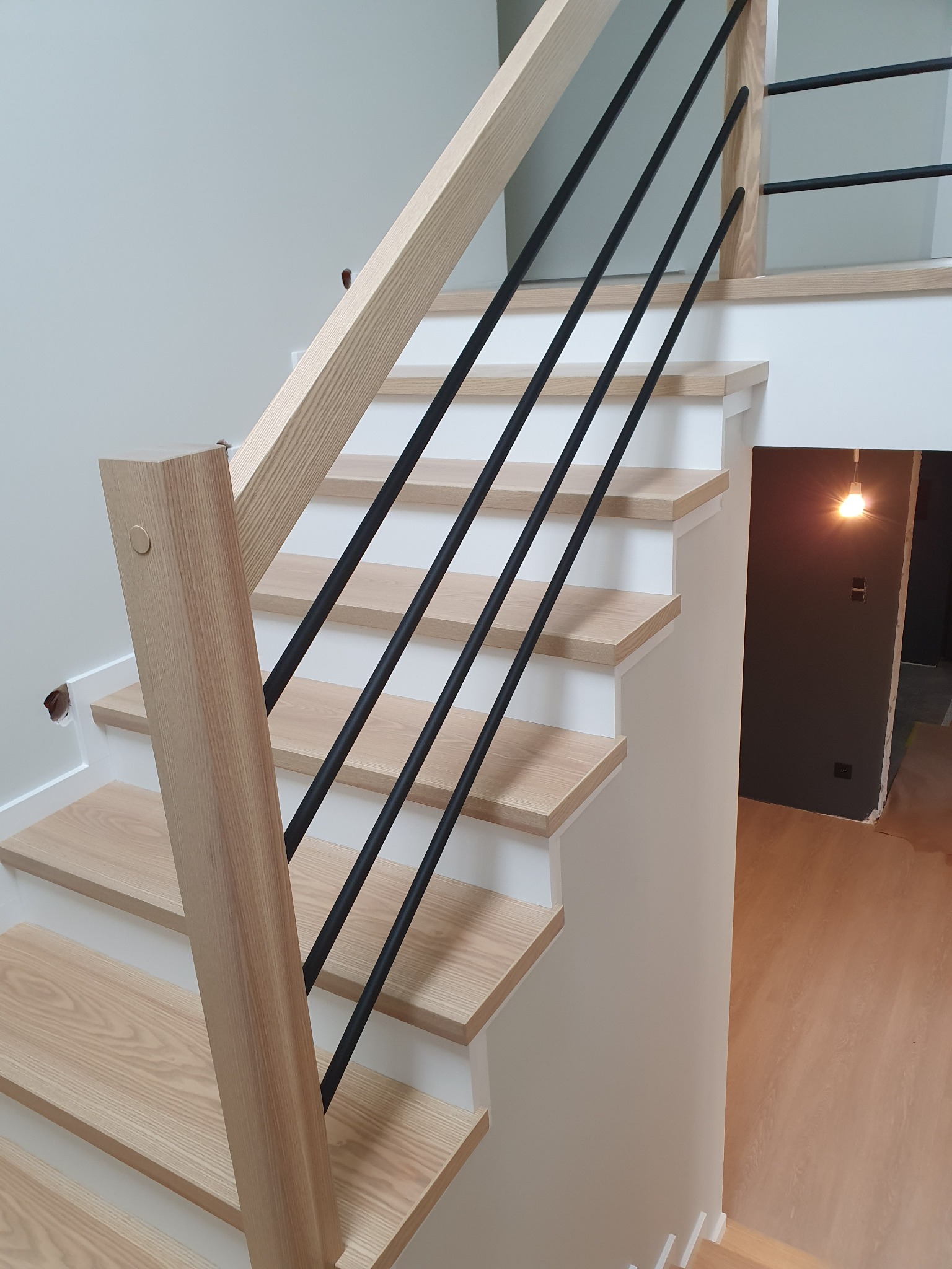 Jasne drewniane stopnie z czarnymi, metalowymi balustradami i drewnianymi słupkami, widoczne niedokończone prace wykończeniowe przy ścianie i podłodze w tle.