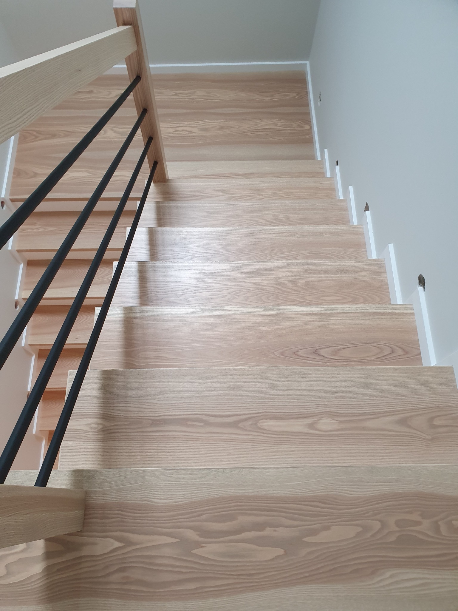 Widok z góry na jasne, drewniane schody z czarnymi, metalowymi balustradami, oświetlone naturalnym światłem, ukazujące wyraźny rysunek słojów drewna.