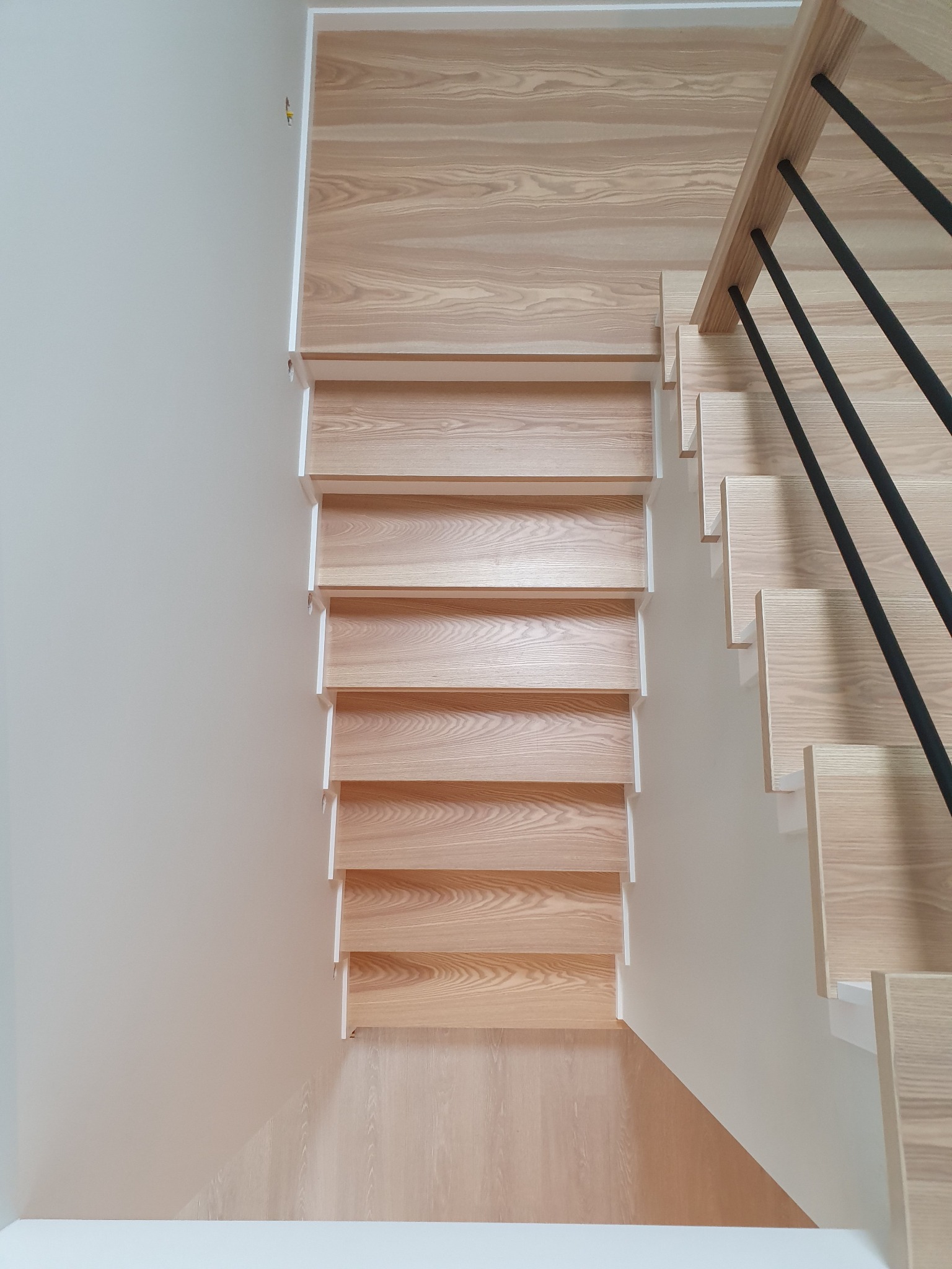 Widok z góry na jasne, drewniane schody z czarną balustradą, przylegające do białej ściany.