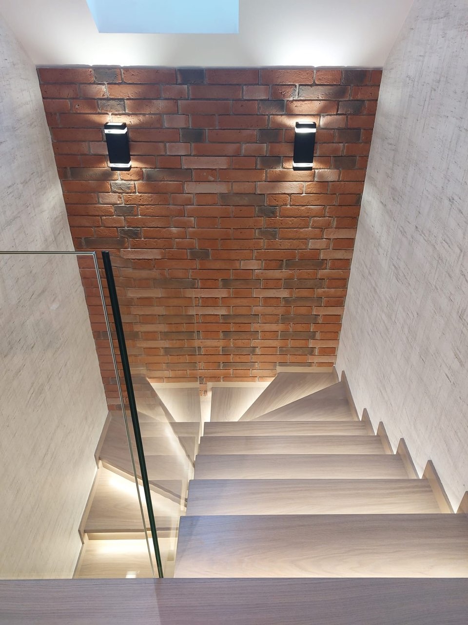Nowoczesne schody z jasnego drewna z podświetleniem LED, szklaną balustradą i ścianą z czerwonej cegły podświetloną czarnymi lampami ściennymi.