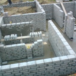 MATEO INTERNATIONAL Sp. z o.o. - Wznoszenie ścian fundamentowych z bloczków betonowych; widoczne fragmenty ścian, zaprawa, drewniane podpory i narzędzia, w tle ziemia i pracownik budowlany.