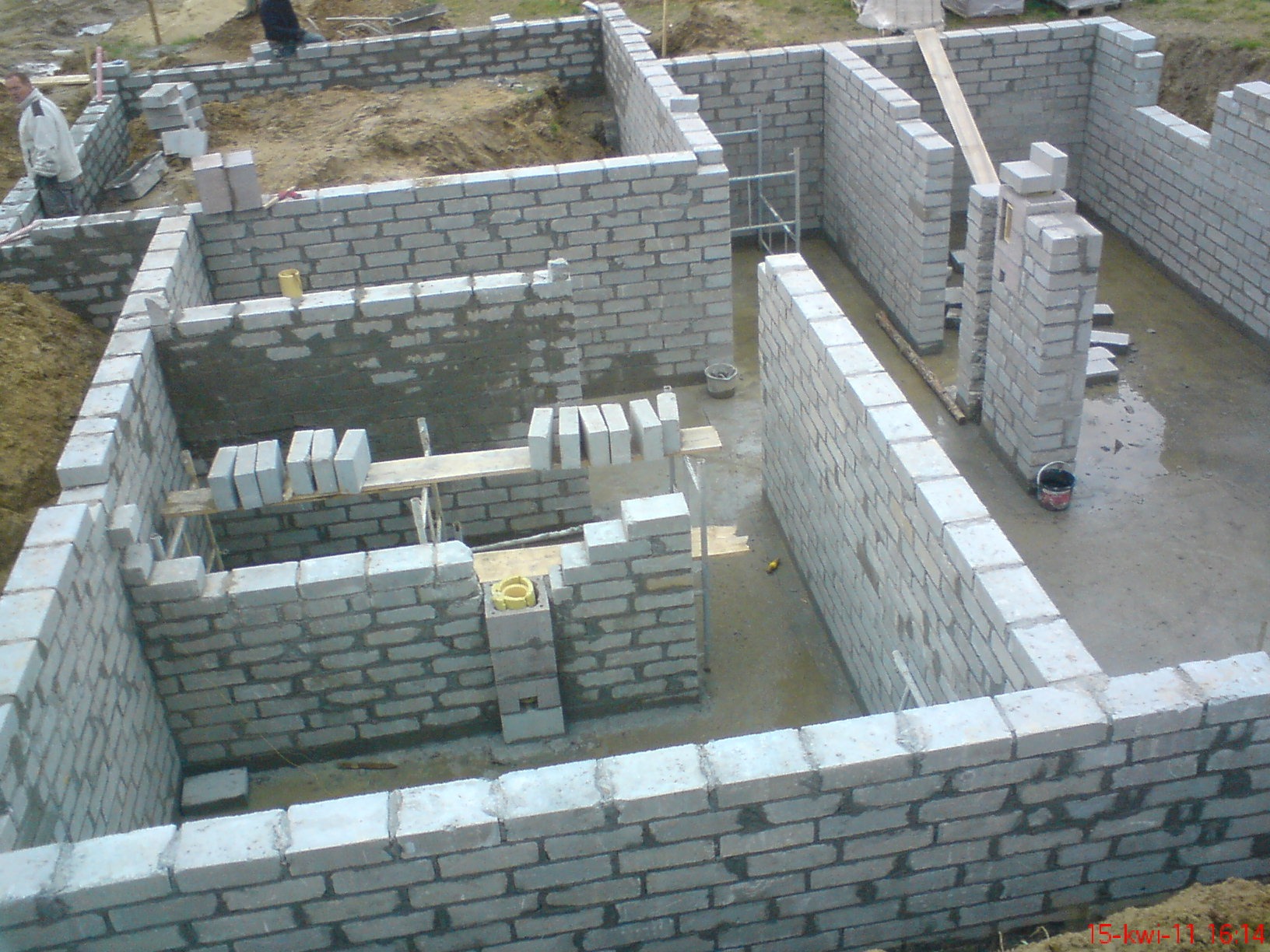 Wznoszenie ścian fundamentowych z bloczków betonowych; widoczne fragmenty ścian, zaprawa, drewniane podpory i narzędzia, w tle ziemia i pracownik budowlany.