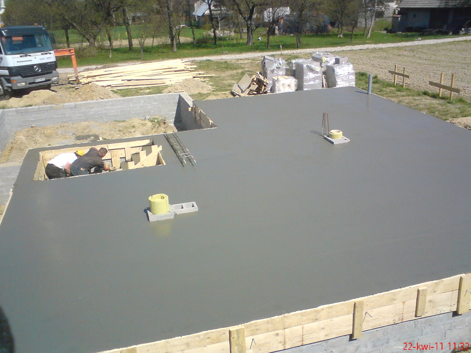 Wylany fundament z deskowaniem i pracownikami przy studzience kanalizacyjnej, widoczne zbrojenia i przygotowane materiały budowlane w tle.