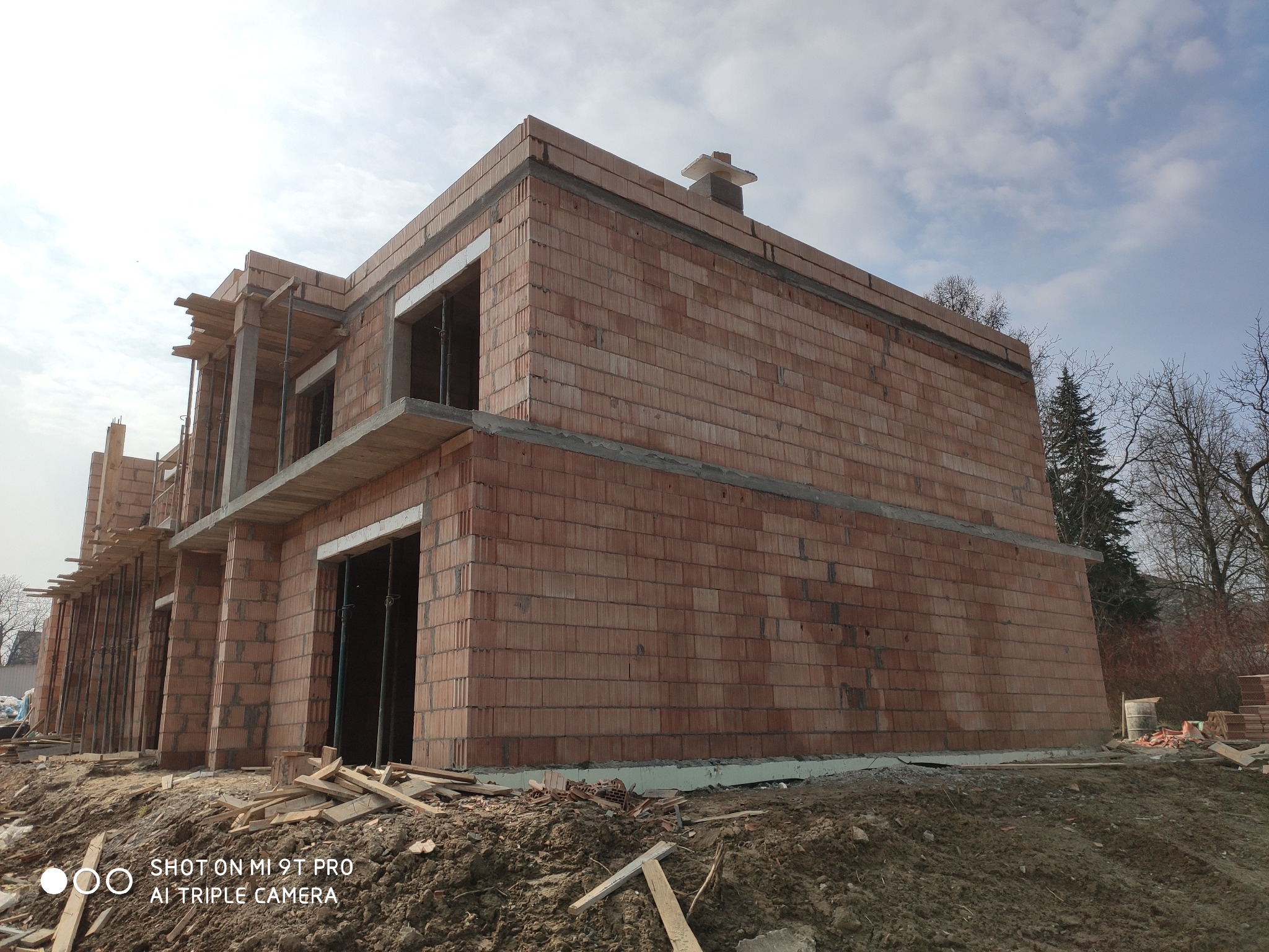 Surowy stan dwukondygnacyjnego budynku z czerwonej cegły ceramicznej, widoczne deskowanie i zbrojenia, teren budowy z rozrzuconymi elementami drewnianymi, pochmurne niebo w tle.