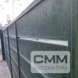 Nowoczesna brama wjazdowa z poziomych paneli w kolorze ciemnej zieleni, pokryta szronem, z widocznym logo firmy 'CMM Automatyka' w dolnym rogu.  Widok z boku, z fragmentem chodnika z kostki brukowej.