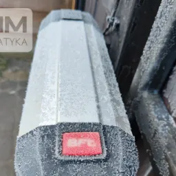 Napęd bramy BFT pokryty szronem, zamontowany na drewnianej bramie, widoczny logotyp CMM Automatyka w tle.