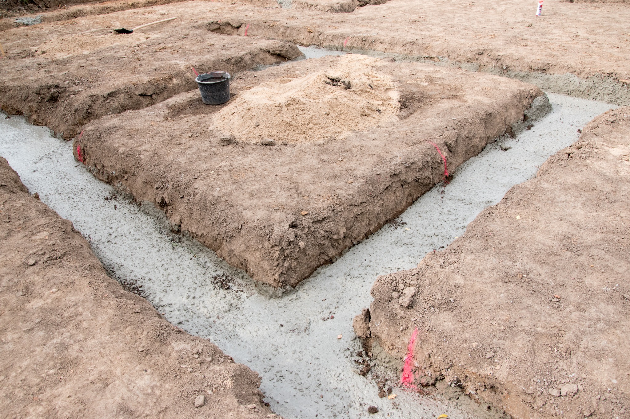 Wylane fundamenty z betonu w wykopach, widoczny kubeł i pryzma piasku na placu budowy.