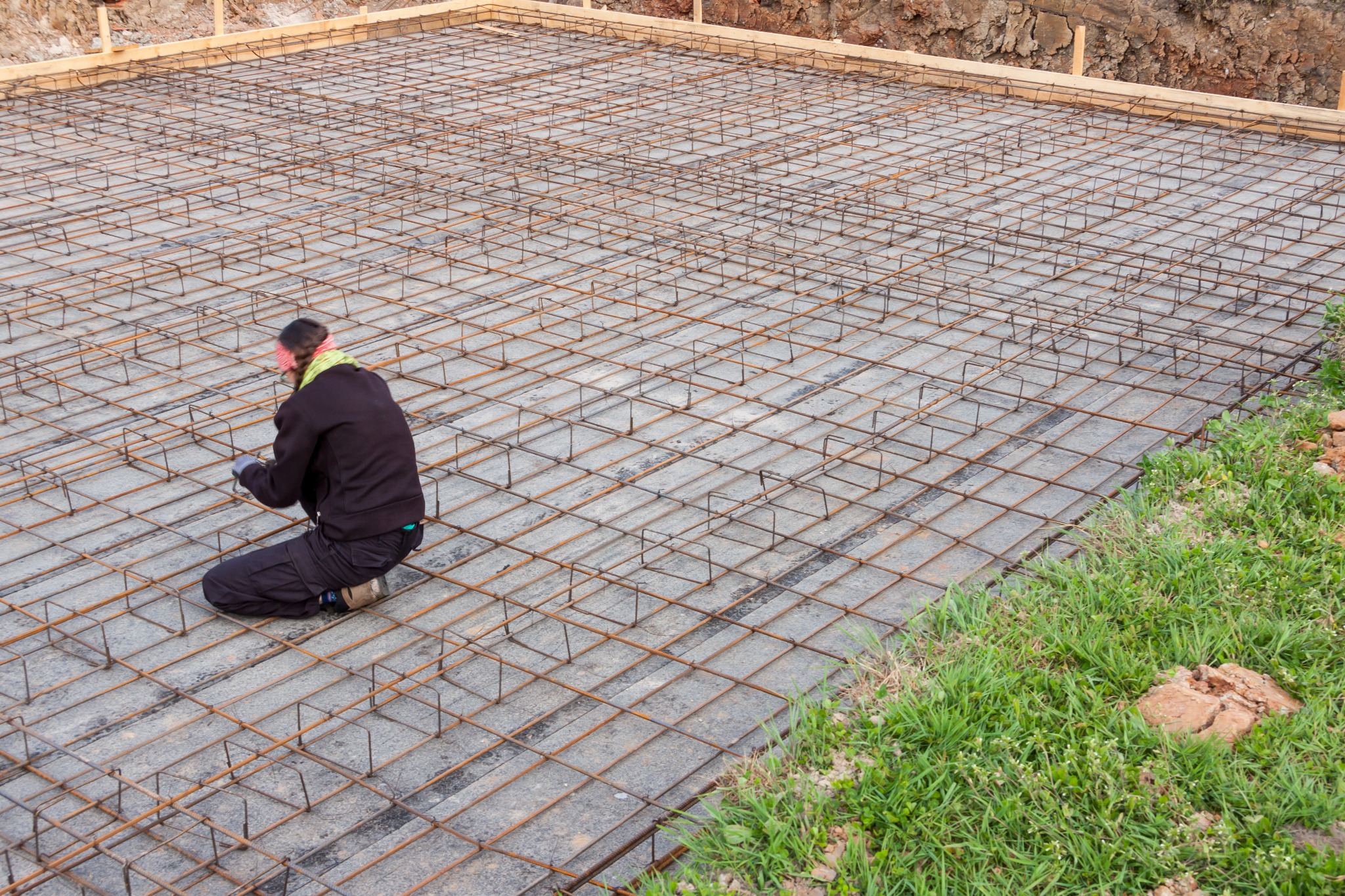 Pracownik w rękawicach wiąże zbrojenie na przygotowanym fundamencie pod budowę, widoczne drewniane szalunki i fragment wykopu.