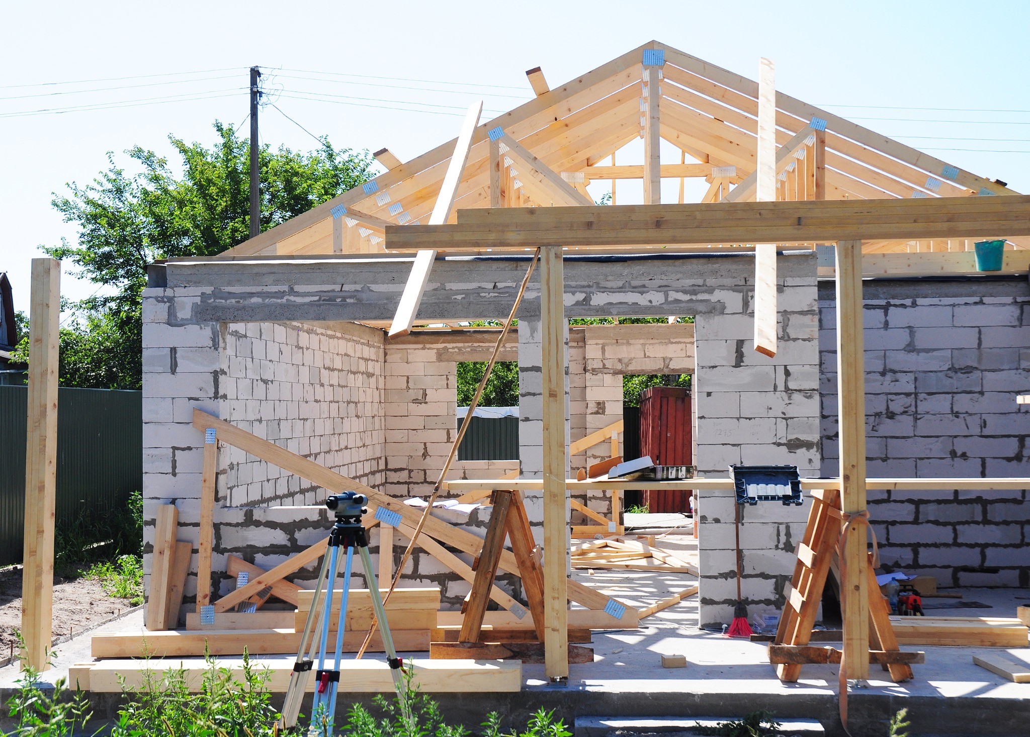Wznoszenie ścian z betonu komórkowego i konstrukcji drewnianej dachu na budowie domu jednorodzinnego, widoczny statyw geodezyjny i tymczasowe podpory.