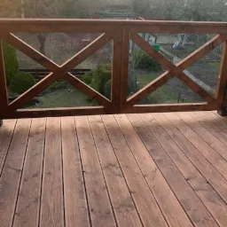 Drewniany taras z brązowymi deskami i balustradą w kształcie krzyży, widok na ogród i drzewa w porannym świetle.