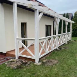 Biała konstrukcja z drewnianą balustradą w kształcie krzyżyków, przylegająca do budynku z brązowymi oknami, widoczne niedokończone prace ziemne wokół podstawy, na ziemi leżą cegły.