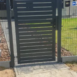 Nowoczesna, grafitowa furtka z poziomymi panelami w ogrodzeniu z siatki, z widocznym monitoringiem na słupku w tle.