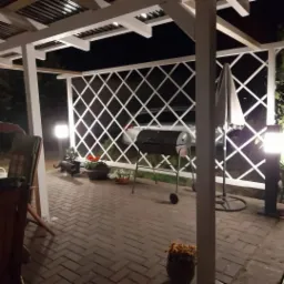 Wieczorny widok na oświetloną altanę z białymi kratownicami, grillem, parasolem i kwiatami w donicach, z kostką brukową na podłożu.