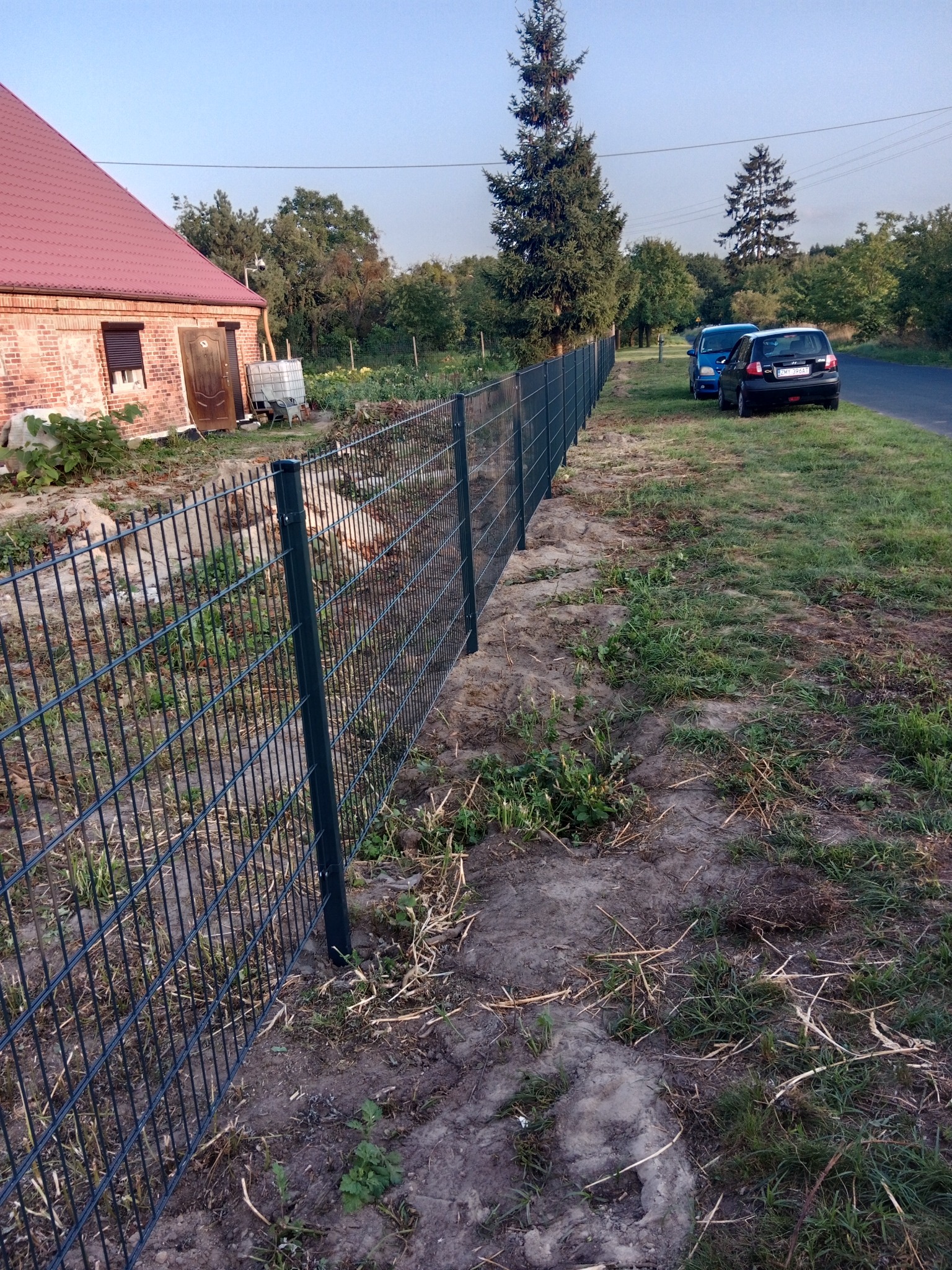 Nowo zainstalowane, metalowe ogrodzenie panelowe w kolorze grafitowym, oddzielające posesję z ceglanym domem od drogi, z zaparkowanymi samochodami w tle.