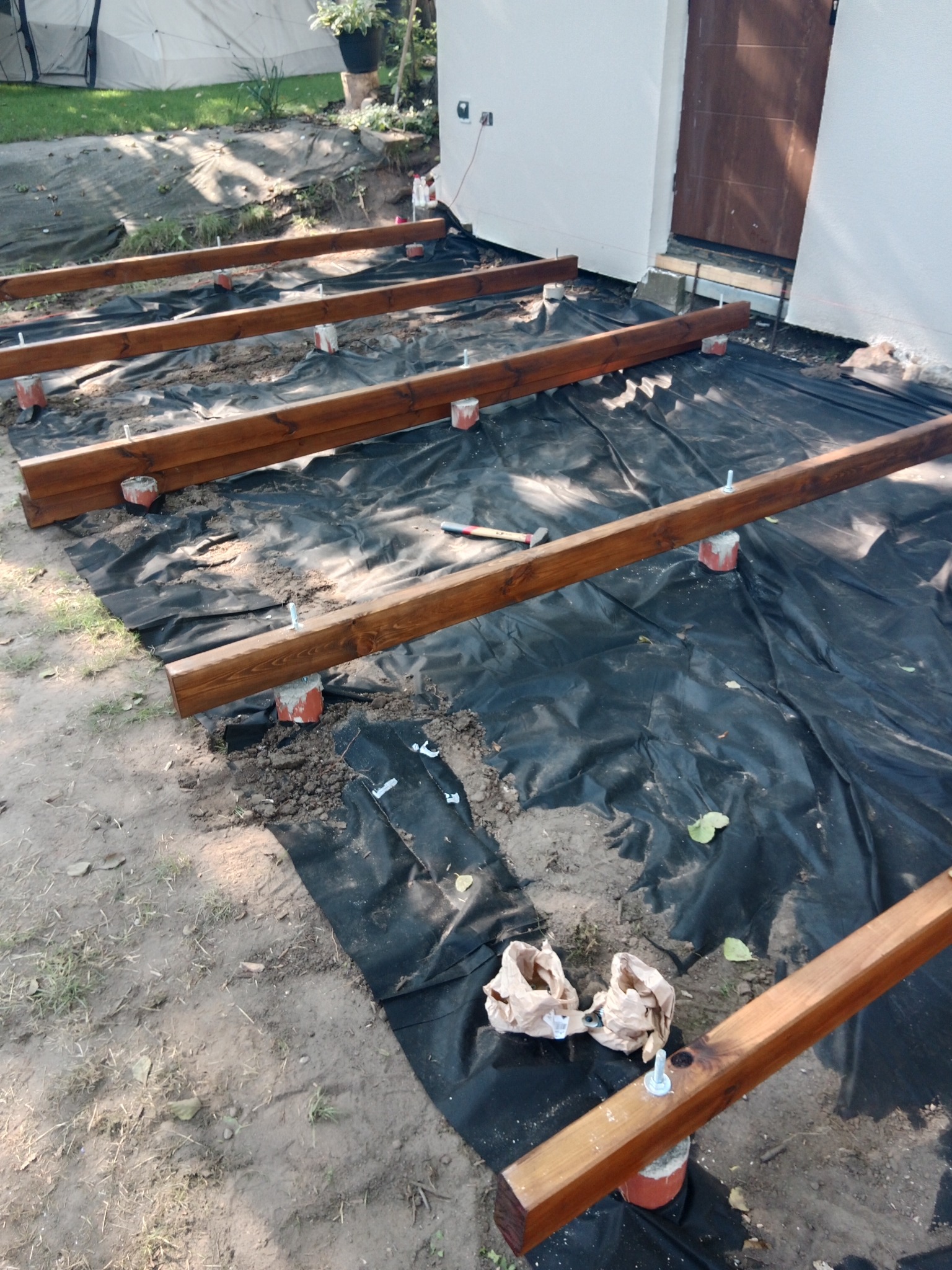 Przygotowanie podłoża pod konstrukcję tarasu: drewniane legary na betonowych wspornikach, rozłożona czarna folia, widoczne narzędzia i materiały budowlane.