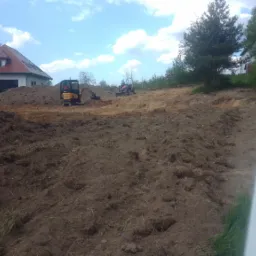 Mini koparka i traktor wyrównują teren pod budowę domu jednorodzinnego, widoczne świeże pryzmy ziemi i ślady opon.