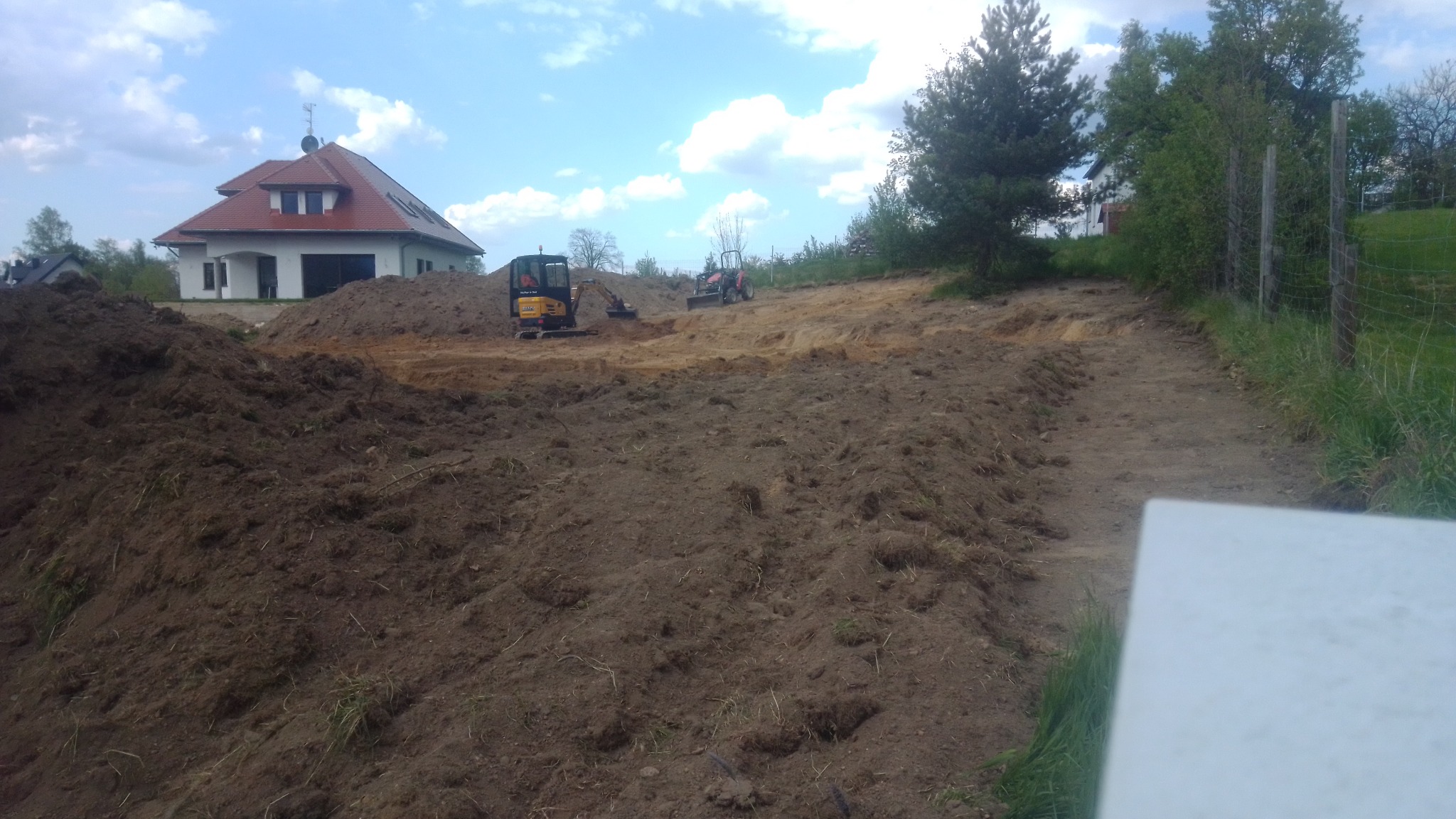 Mini koparka i traktor wyrównują teren pod budowę domu jednorodzinnego, widoczne świeże pryzmy ziemi i ślady opon.