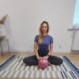 Kobieta w okularach siedzi na podłodze na wzorzystym dywanie, trzymając małą, różową piłkę do ćwiczeń; w tle ściana z ramkami.