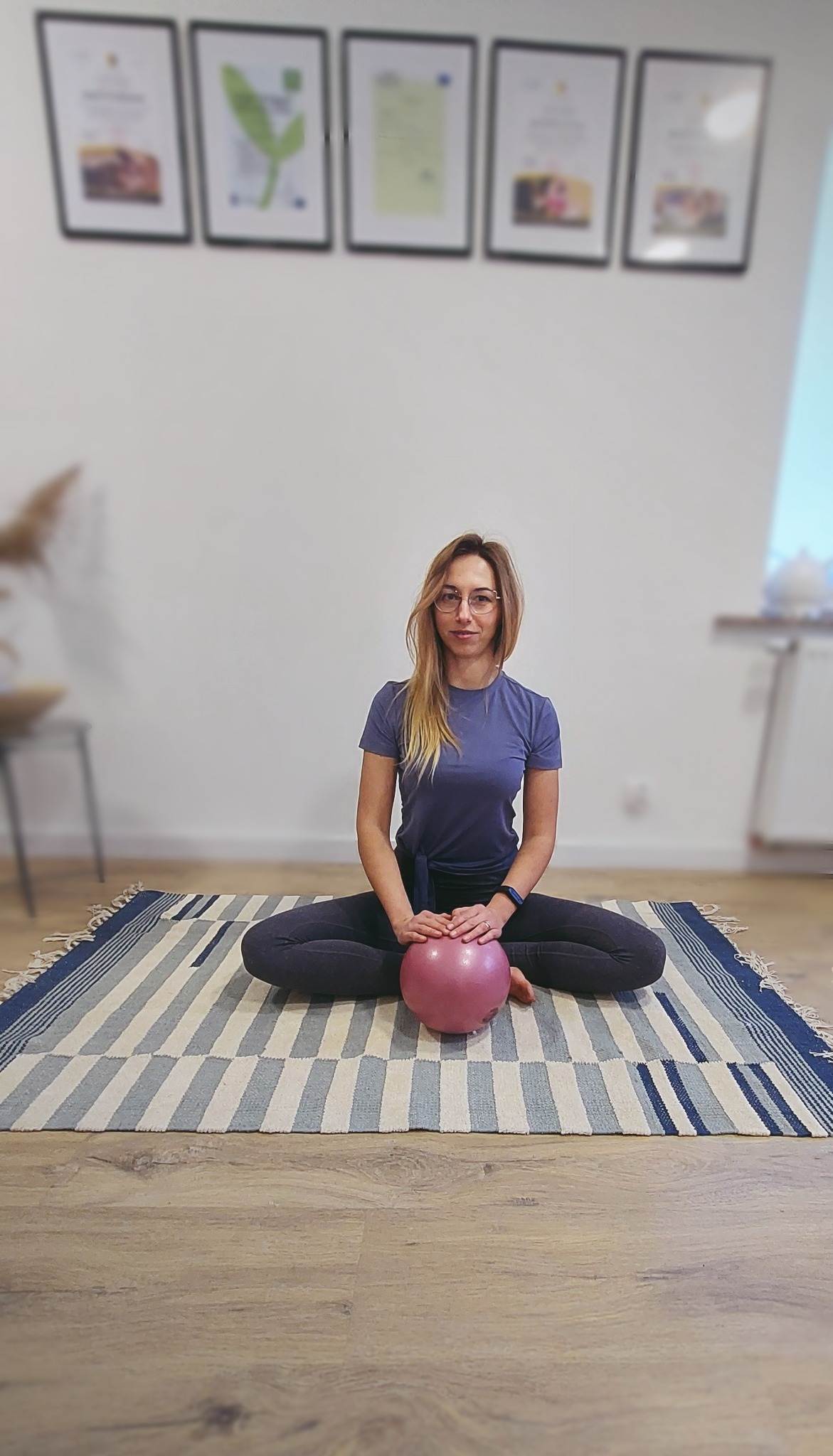 Kobieta w okularach siedzi na podłodze na wzorzystym dywanie, trzymając małą, różową piłkę do ćwiczeń; w tle ściana z ramkami.