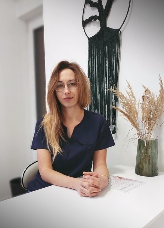 Kobieta w okularach i granatowym uniformie siedzi przy białym biurku w gabinecie, za nią wazon z suszonymi trawami i makrama w kształcie drzewa.