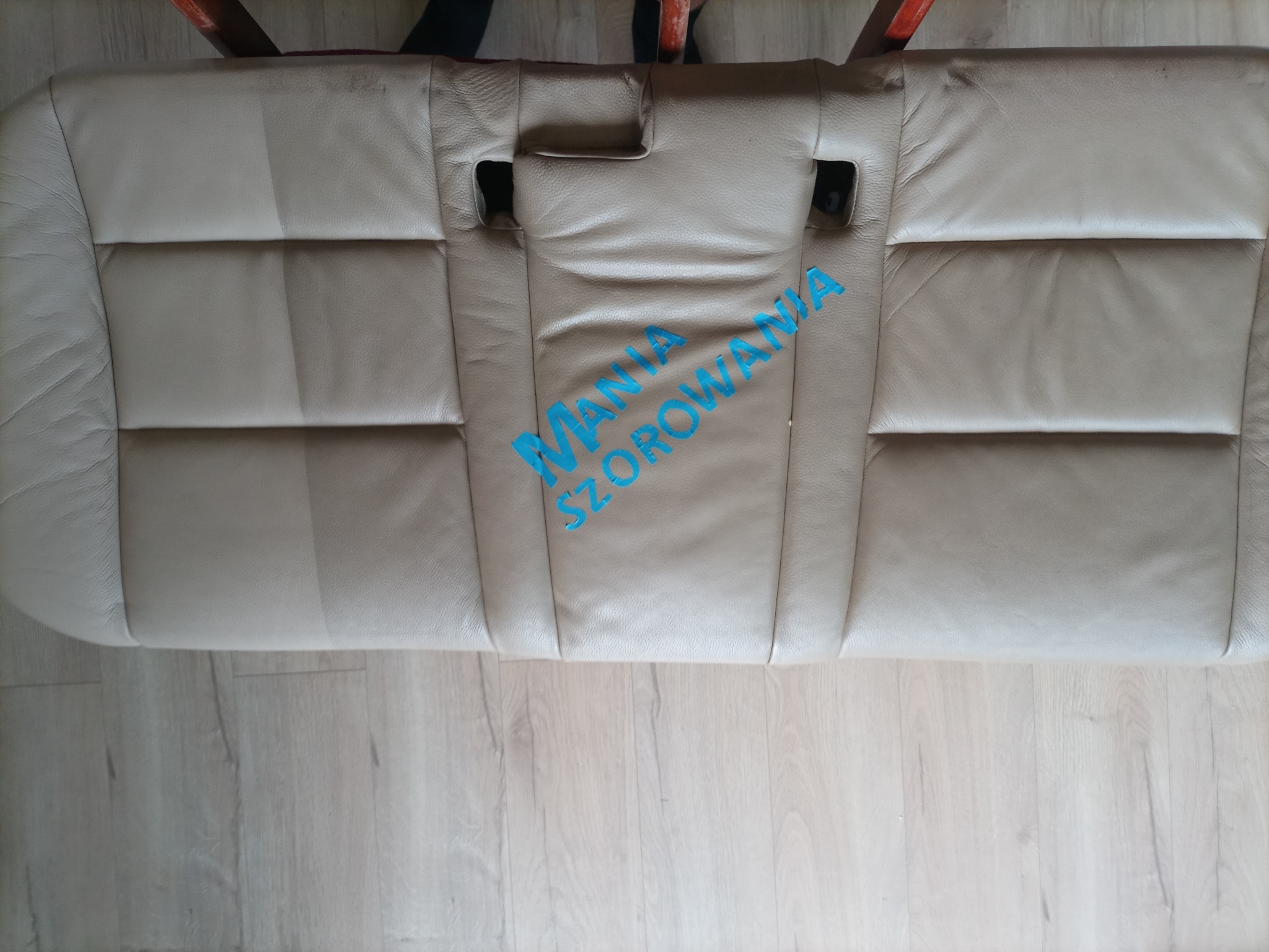 Beżowa skórzana tylna kanapa samochodowa z widoczną różnicą w czystości po prawej stronie, z napisem 'Mania Szorowania' naniesionym cyfrowo, leżąca na podłodze z paneli.