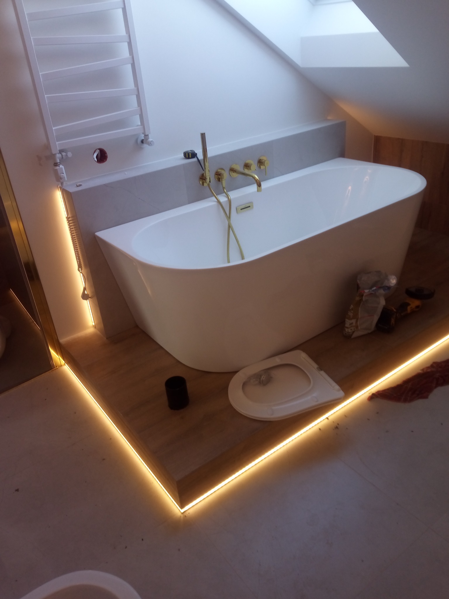 Nowoczesna łazienka na poddaszu z wolnostojącą wanną, złotą armaturą i podświetleniem LED. Widoczne narzędzia i akcesoria montażowe.