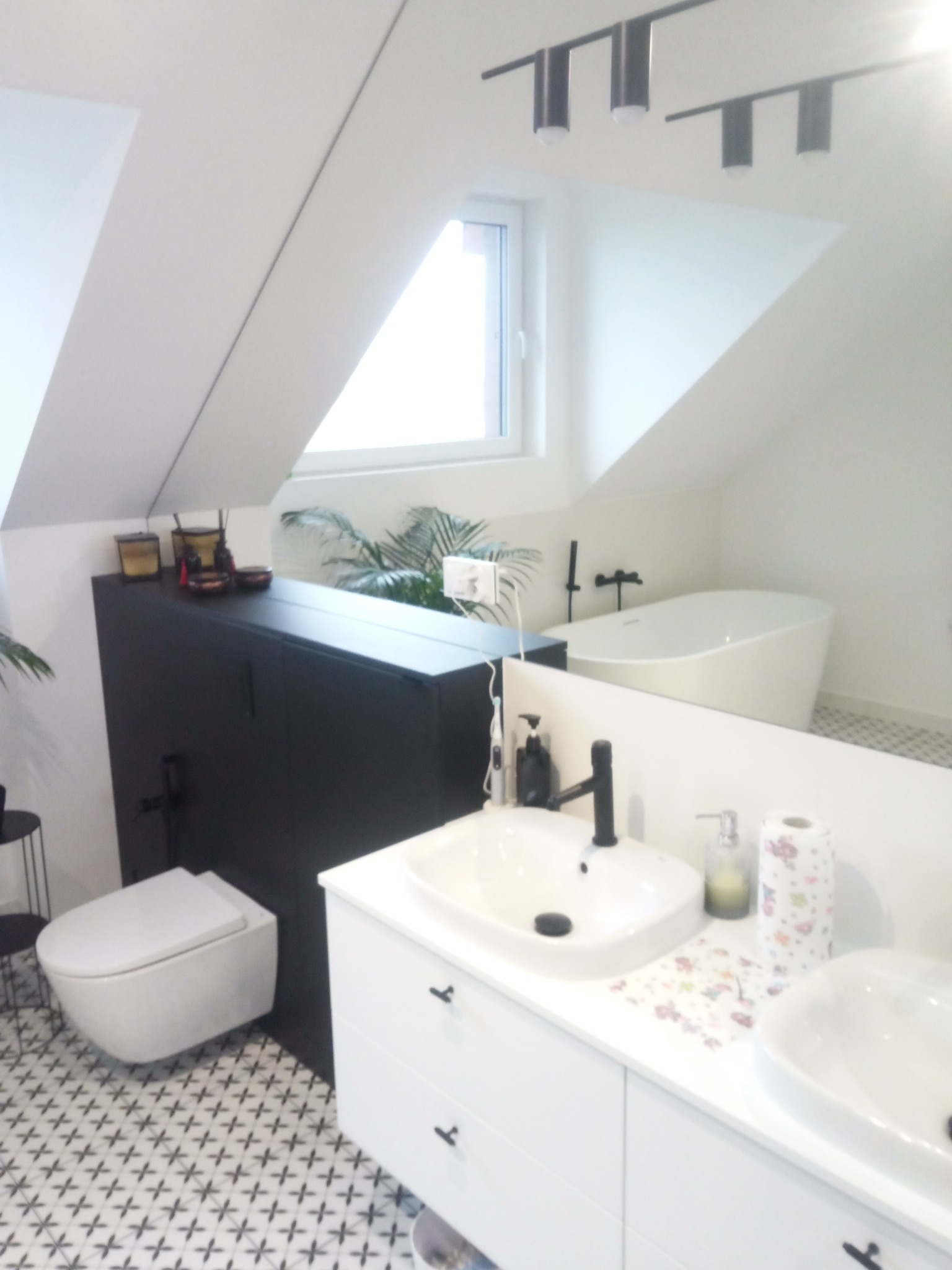 Jasna łazienka na poddaszu z wanną, umywalką i toaletą. Uwagę przykuwają czarne akcenty: bateria, szafka i oświetlenie. Na blacie dekoracyjny dozownik i ręcznik papierowy.