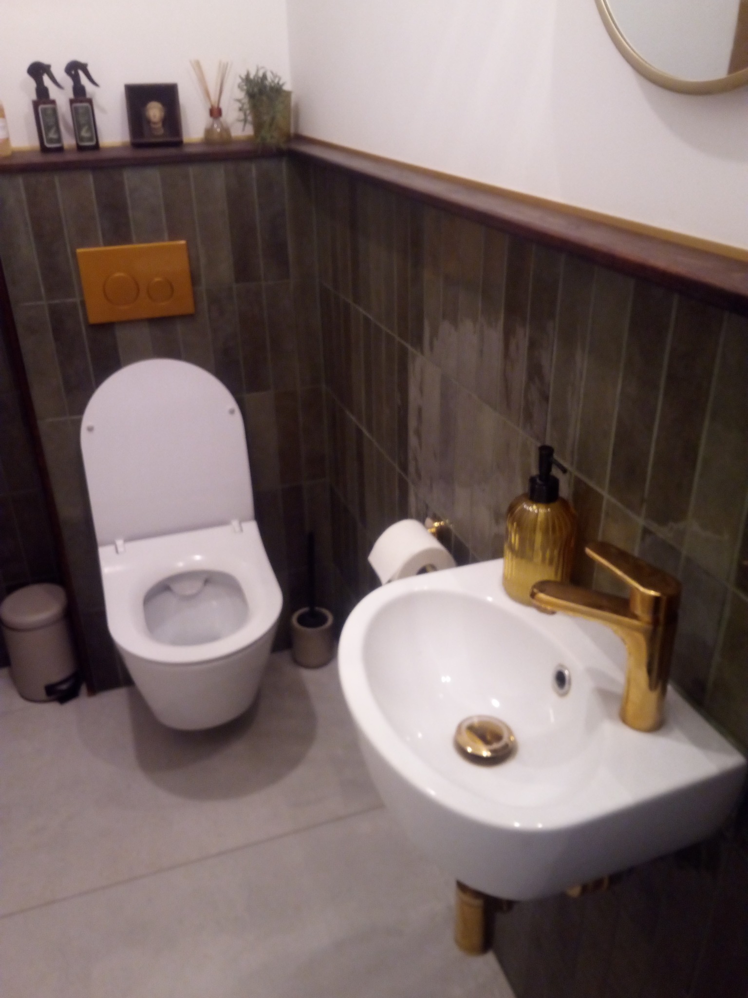 Nowoczesna łazienka z wiszącą toaletą i umywalką z baterią w kolorze złota, wykończona ciemnymi płytkami. Widoczne akcesoria łazienkowe i dekoracje na półce.