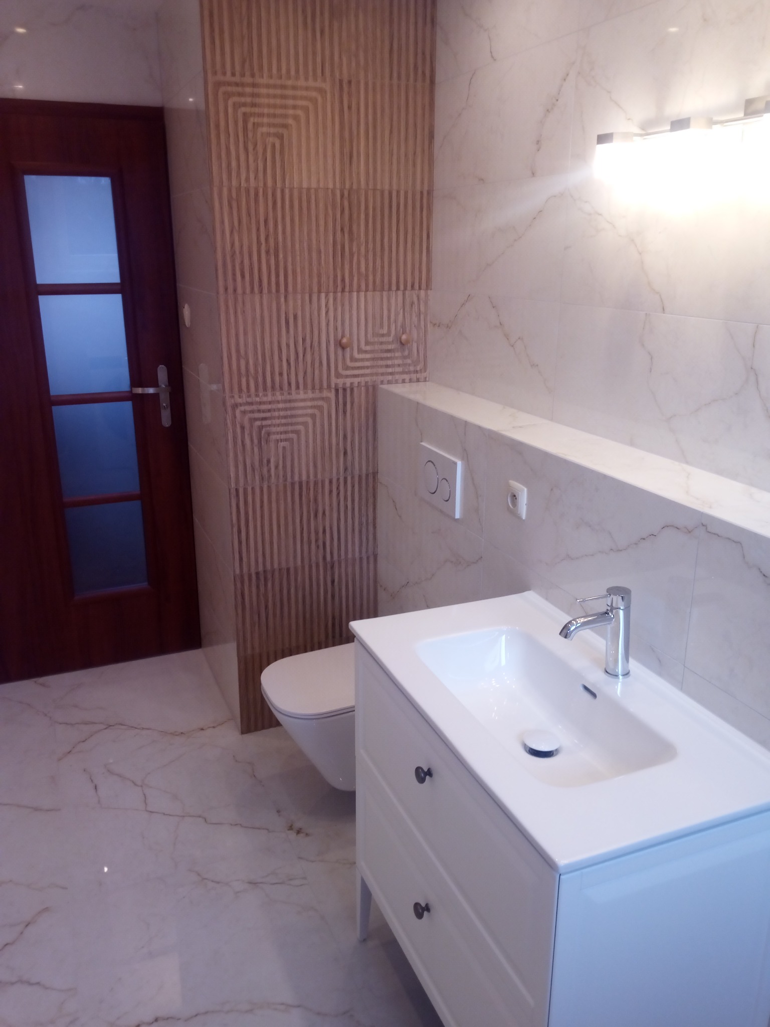 Nowoczesna łazienka po remoncie: biała umywalka z szafką, toaleta, marmurowe płytki i dekoracyjna ściana z drewnianymi panelami. Oświetlenie górne.