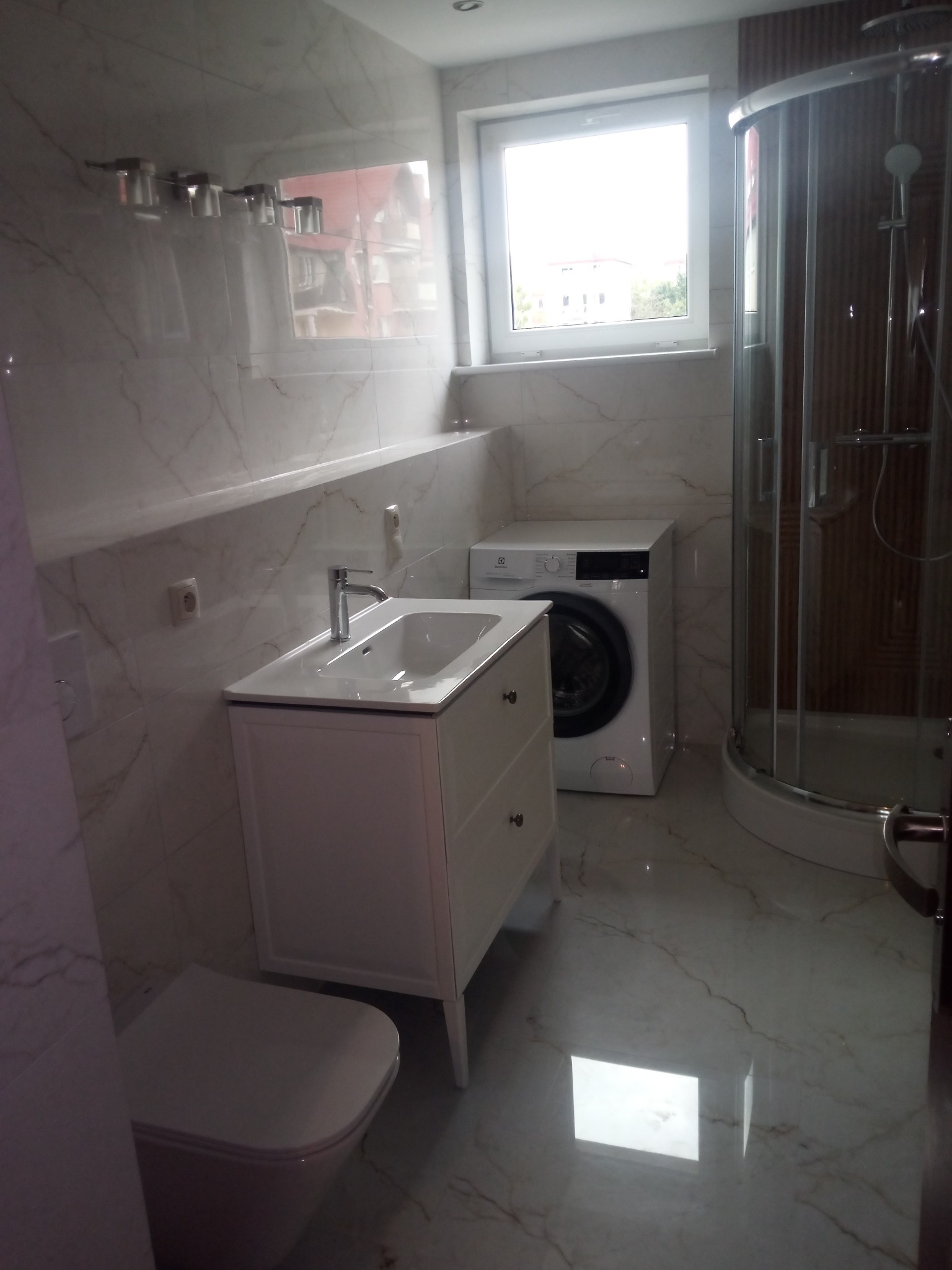 Wykończona łazienka z marmurowymi płytkami, białą szafką z umywalką, toaletą, pralką i kabiną prysznicową. Widok na okno z widokiem na budynek.