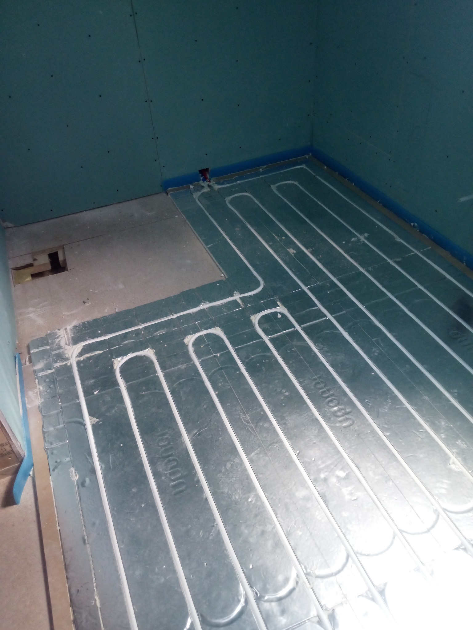 Instalacja ogrzewania podłogowego w łazience, rury ułożone na srebrnej folii izolacyjnej, ściany wykończone płytami gipsowo-kartonowymi, widoczny fragment podłogi bez izolacji.