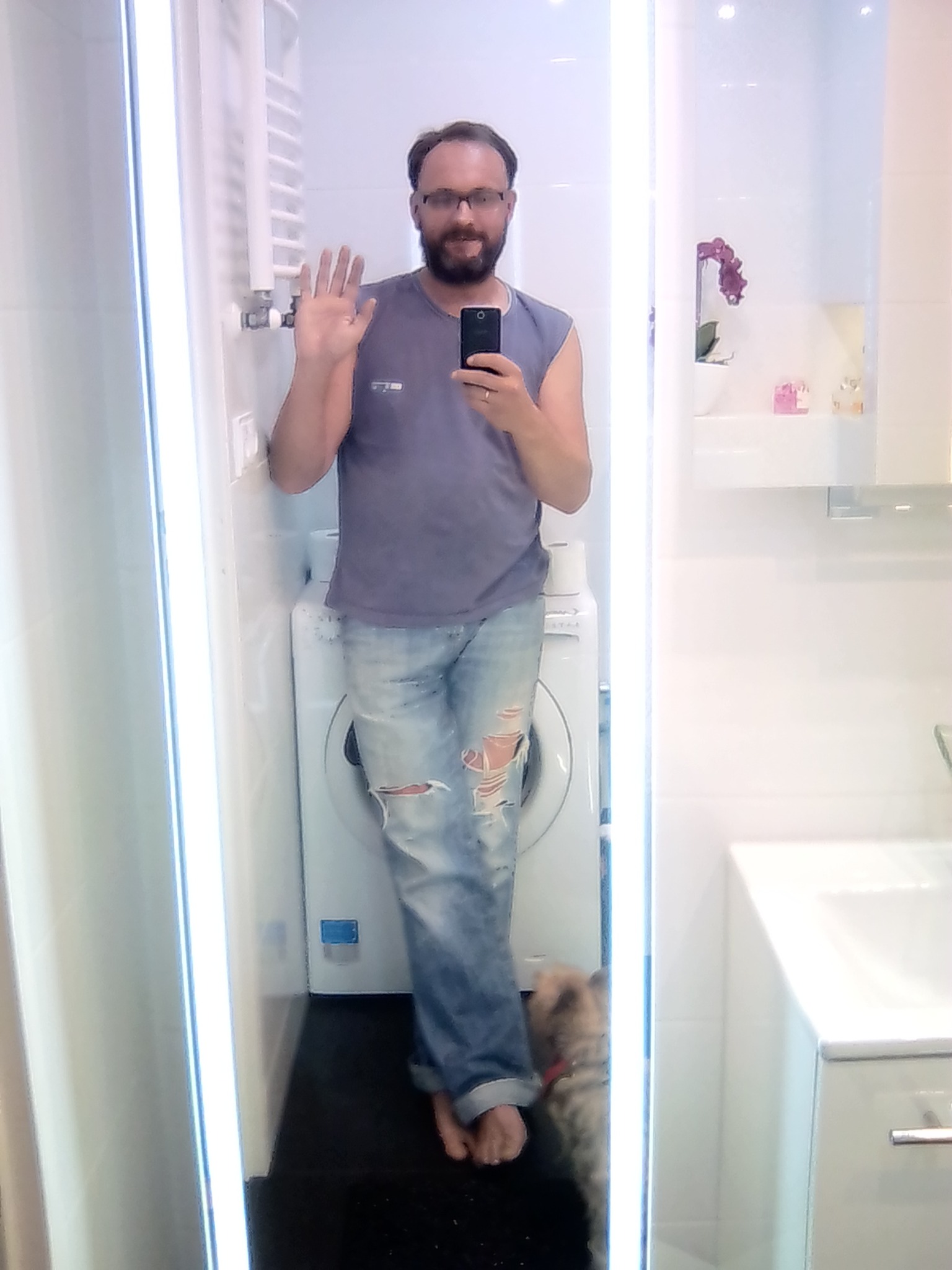 Mężczyzna robi selfie w łazience z pralką w tle, uśmiechnięty i machający ręką, widoczny pies. Oświetlenie LED w lustrze.