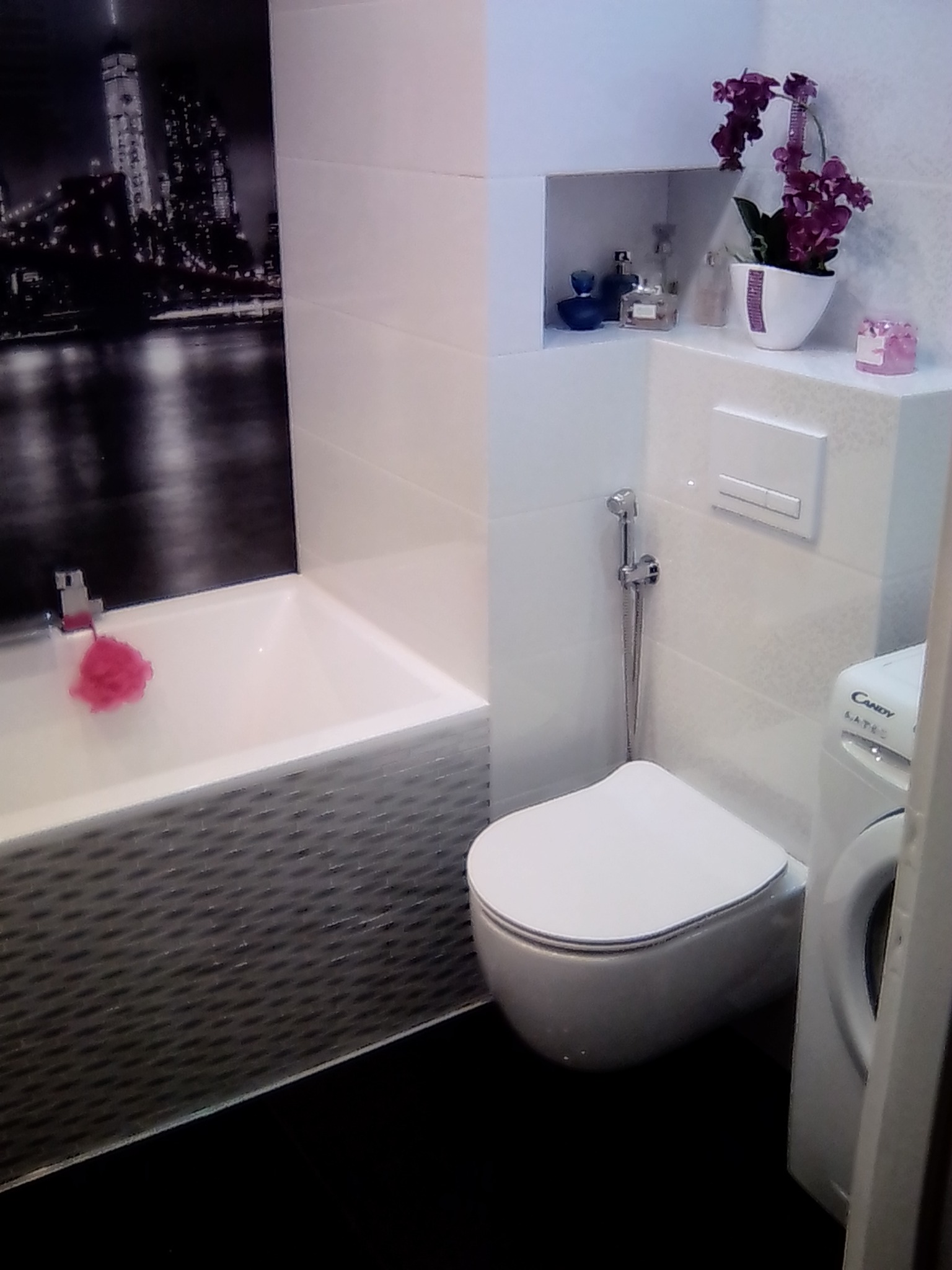 Nowoczesna łazienka z wanną, toaletą i pralką. Na ścianie dekoracyjny panel z motywem miasta. Umywalka z kwiatami i kosmetykami. Ciemna podłoga.